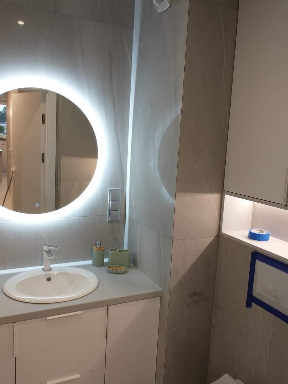 Nowoczesna łazienka z okrągłym lustrem z podświetleniem LED, szarą armaturą i płytkami imitującymi marmur, oraz białymi szafkami pod umywalką i wiszącymi, z akcesoriami łazienkowymi w odcieniach...