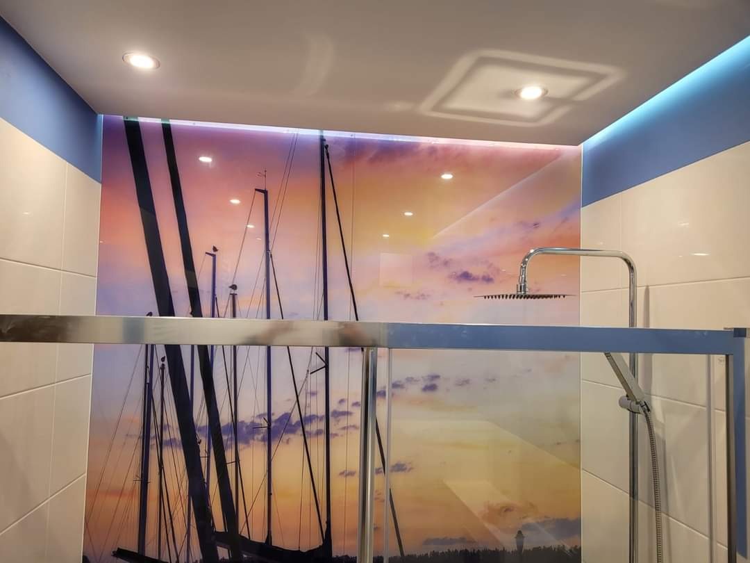 Nowoczesna kabina prysznicowa ze szklaną ścianą ozdobioną motywem żaglówek na tle zachodzącego słońca, biało-niebieskie płytki na ścianach, oświetlenie sufitowe.