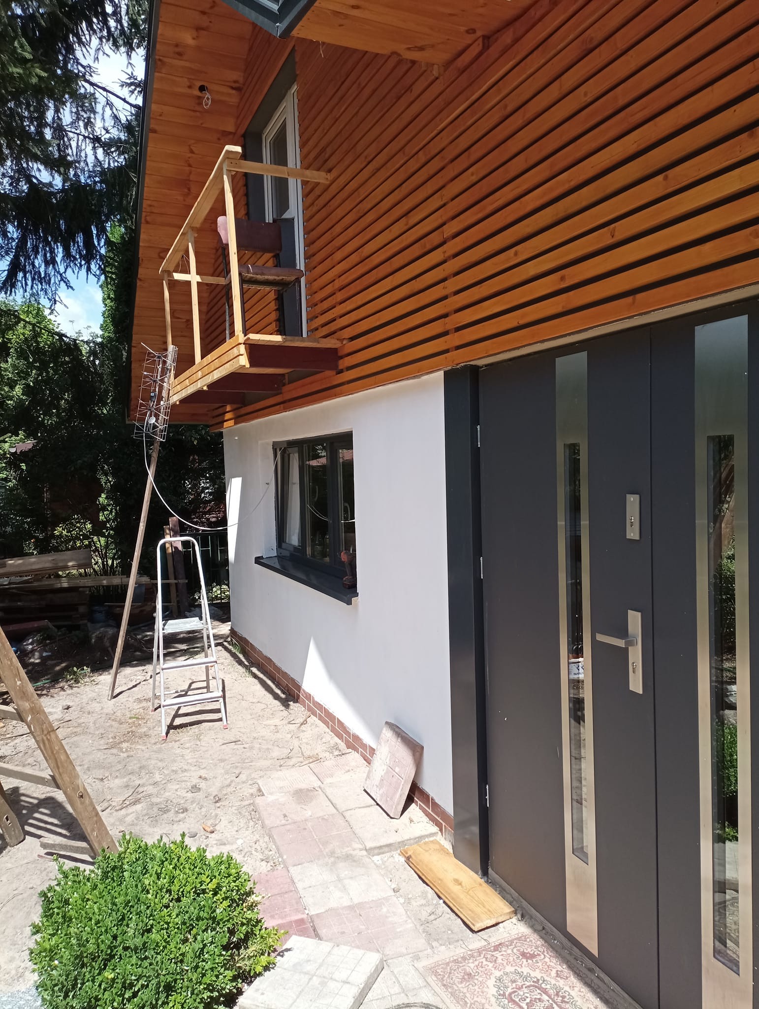 Elewacja domu z drewnianą okładziną, montaż anteny na prowizorycznym balkonie, biała ściana, szare drzwi wejściowe i okno, drabina i deski na ziemi.
