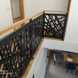 Nowoczesna balustrada schodowa z czarnego metalu o geometrycznym wzorze, uzupełniona jasnym drewnem, widok z g&oacute;ry na jadalnię z krzesłami w odcieniach szarości, granatu i musztardy.