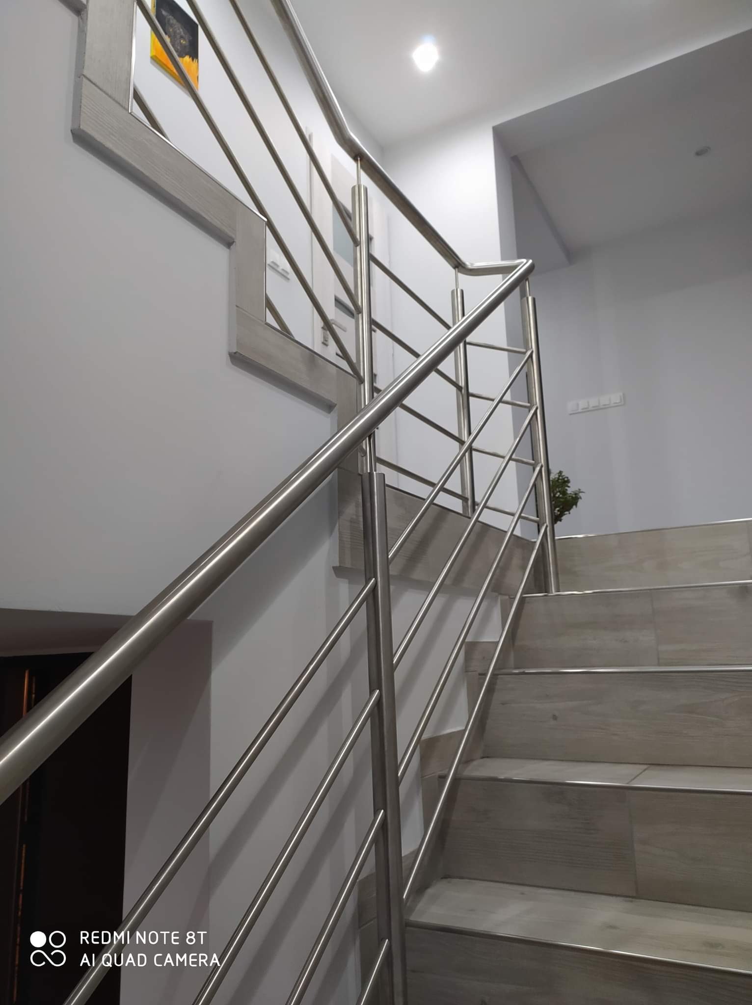 Schody z szarymi płytkami i nowoczesną balustradą ze stali nierdzewnej z poziomymi prętami, widok z dołu, na ścianie dekoracyjny element z obrazkiem.