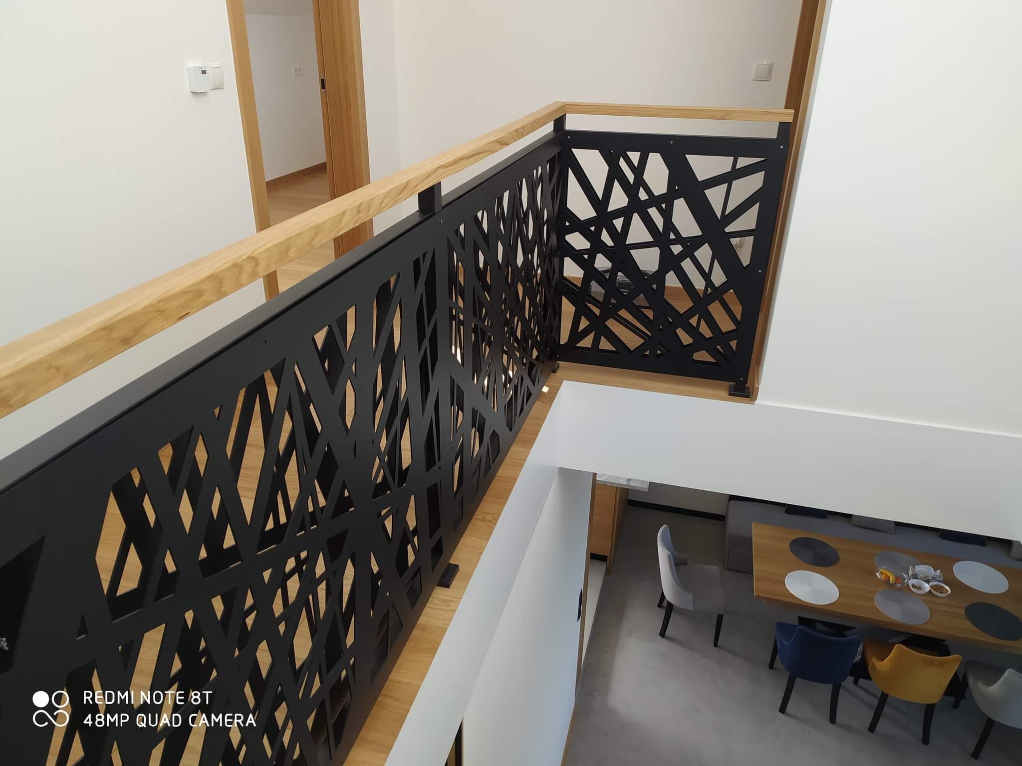 Nowoczesna balustrada schodowa z czarnego metalu o geometrycznym wzorze, uzupełniona jasnym drewnem, widok z góry na jadalnię z krzesłami w odcieniach szarości, granatu i musztardy.