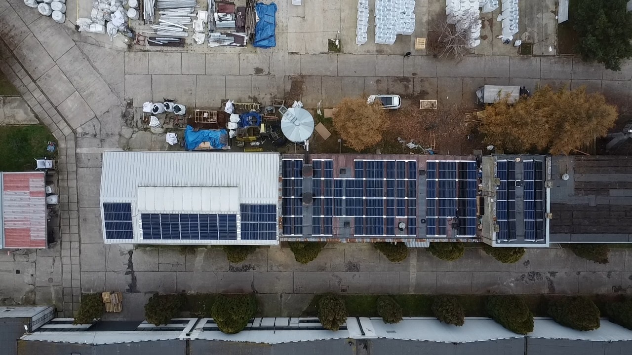 Widok z góry na dachy budynków z zainstalowanymi panelami słonecznymi, widoczny teren wokół budynków, w tle zaparkowany samochód i składowane materiały.