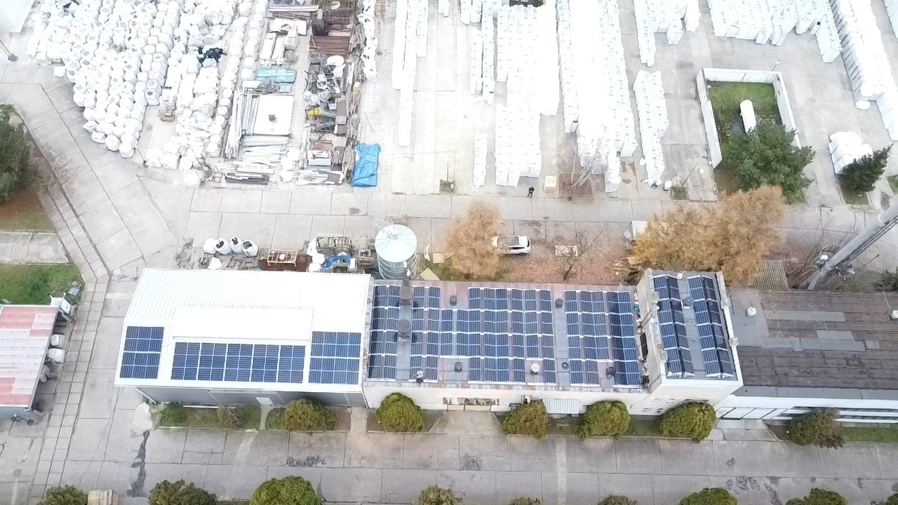 Widok z lotu ptaka na budynek przemysłowy z zainstalowanymi panelami fotowoltaicznymi na dachu, otoczony placem składowym z materiałami budowlanymi i drzewami.