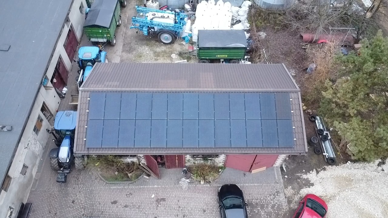 Widok z góry na budynek gospodarczy z brązowym dachem, na którym zamontowane są panele słoneczne, obok stoją traktory i zaparkowane samochody.
