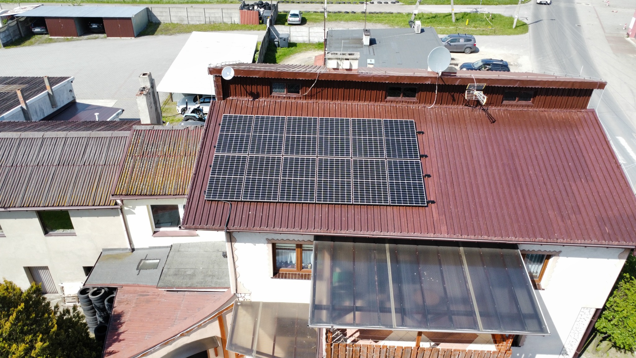 Panel słoneczny na dachu domu z brązową blachodachówką, widok z góry, na drugim planie budynki gospodarcze i droga.