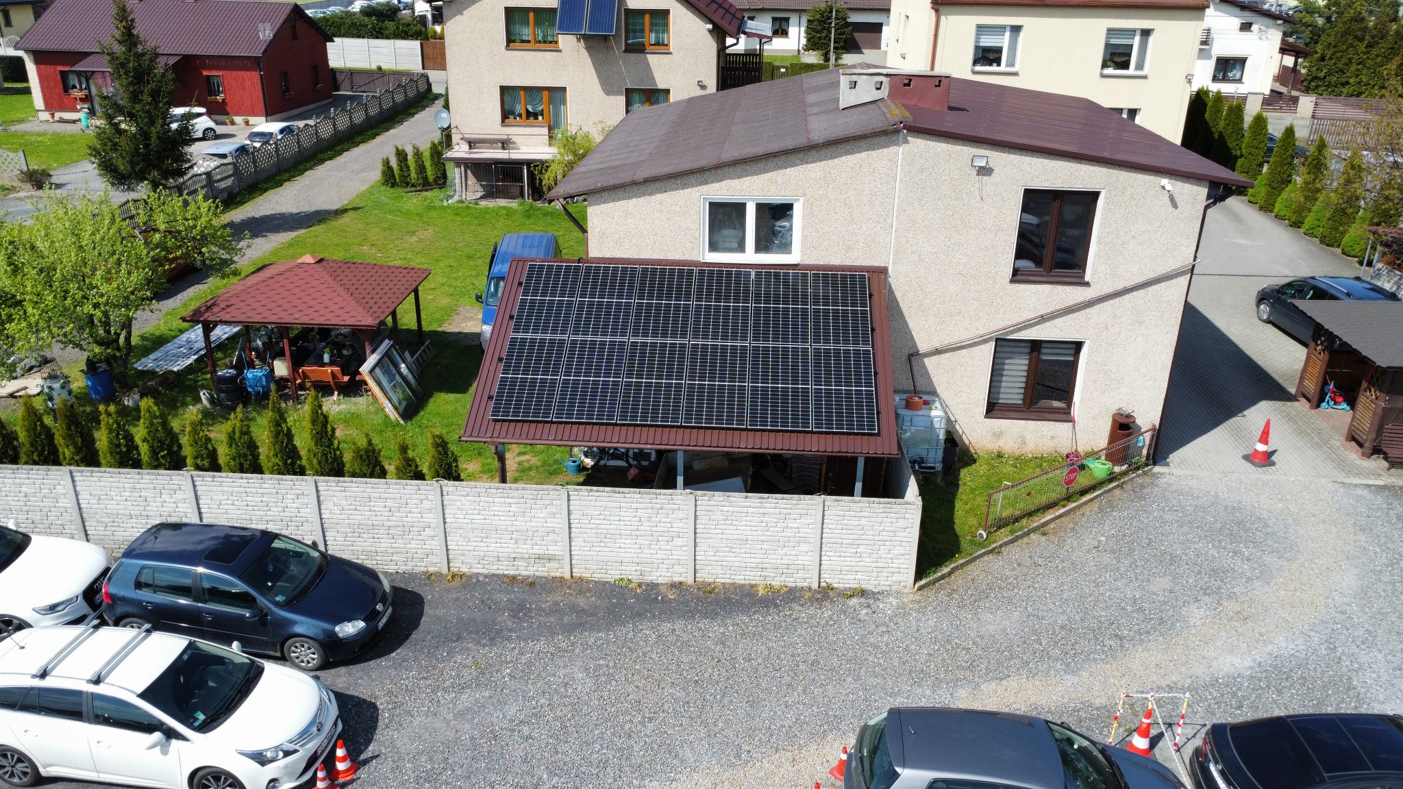 Widok z góry na dom jednorodzinny z panelami słonecznymi zamontowanymi na dachu zadaszenia nad tarasem, widoczne zaparkowane samochody na żwirowym podjeździe i fragment ogrodu z altaną.