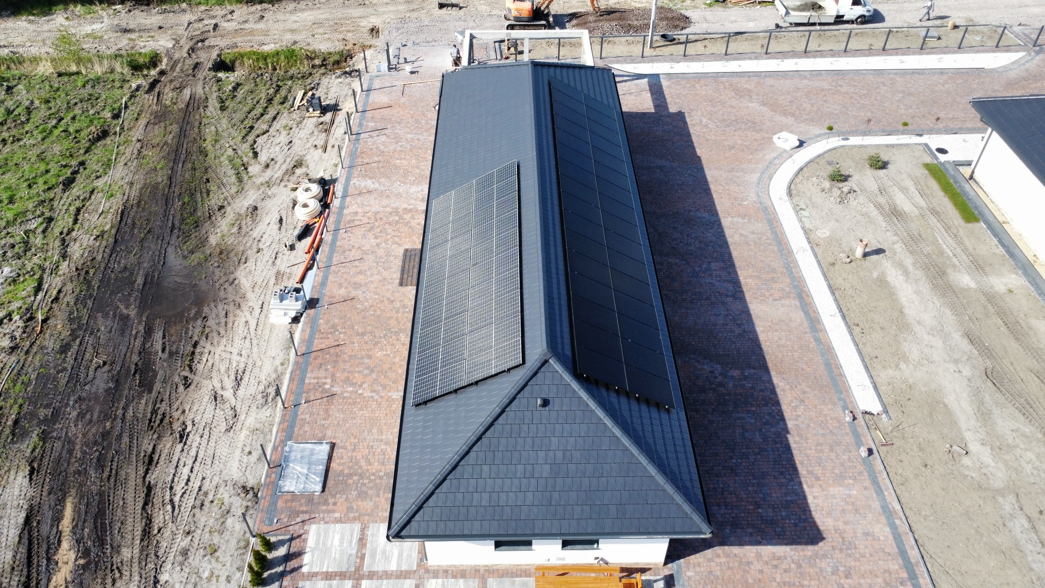 Widok z góry na dach domu z dwoma różnymi zestawami paneli słonecznych, po lewej jasnoszare, po prawej ciemnoszare, otoczonego nową kostką brukową i terenem w trakcie zagospodarowywania.