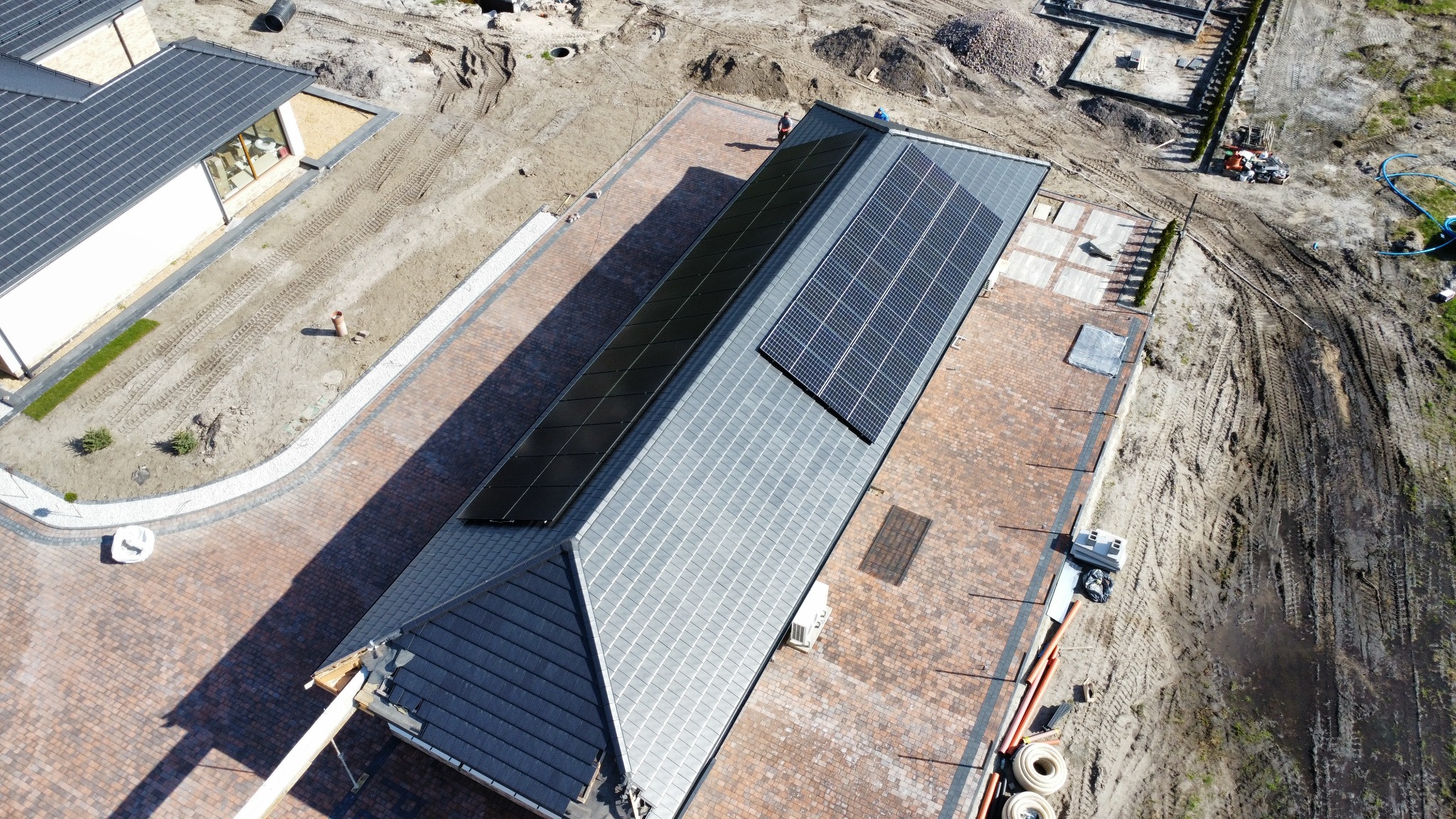 Widok z drona na dach domu z zainstalowanymi panelami fotowoltaicznymi, część paneli w czarnej ramie, część w niebieskiej, na dachu dwóch pracowników podczas montażu, kostka brukowa przed domem.