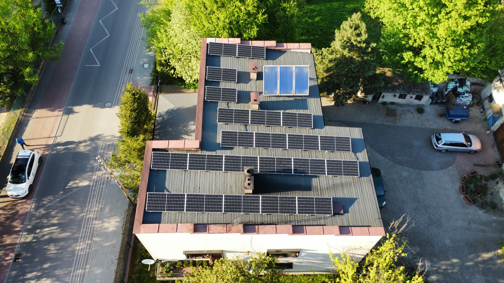 Widok z drona na dach budynku z zainstalowanymi panelami słonecznymi; na dachu widoczny komin, a obok budynku zaparkowany samochód.