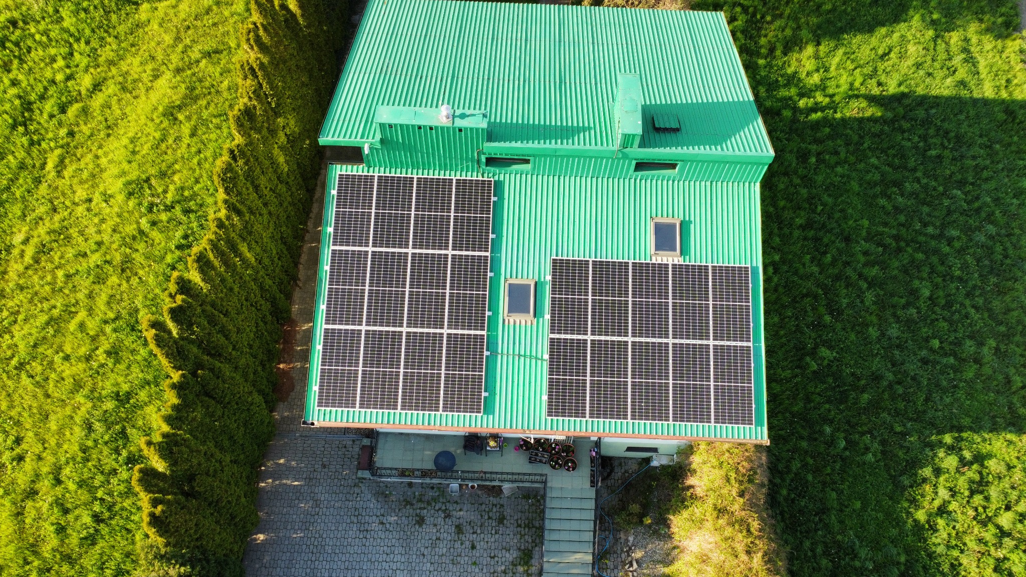 Widok z góry na dom z zielonym dachem z blachy falistej i panelami fotowoltaicznymi, otoczony zielonymi krzewami i trawnikiem.