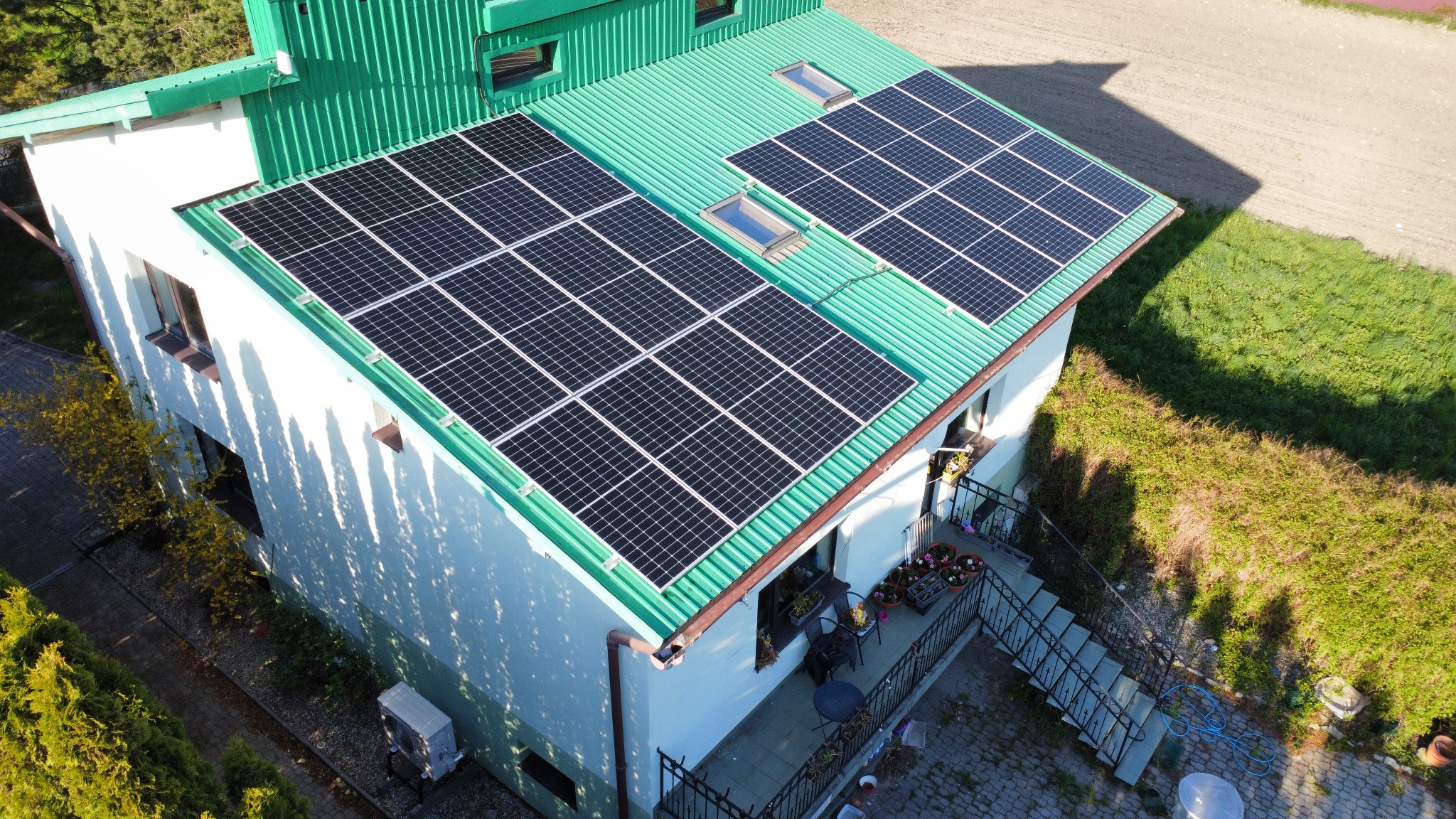 Widok z góry na dom z zielonym dachem z blachy falistej i panelami słonecznymi, z balkonem i schodami oraz zielonym ogrodem w tle.