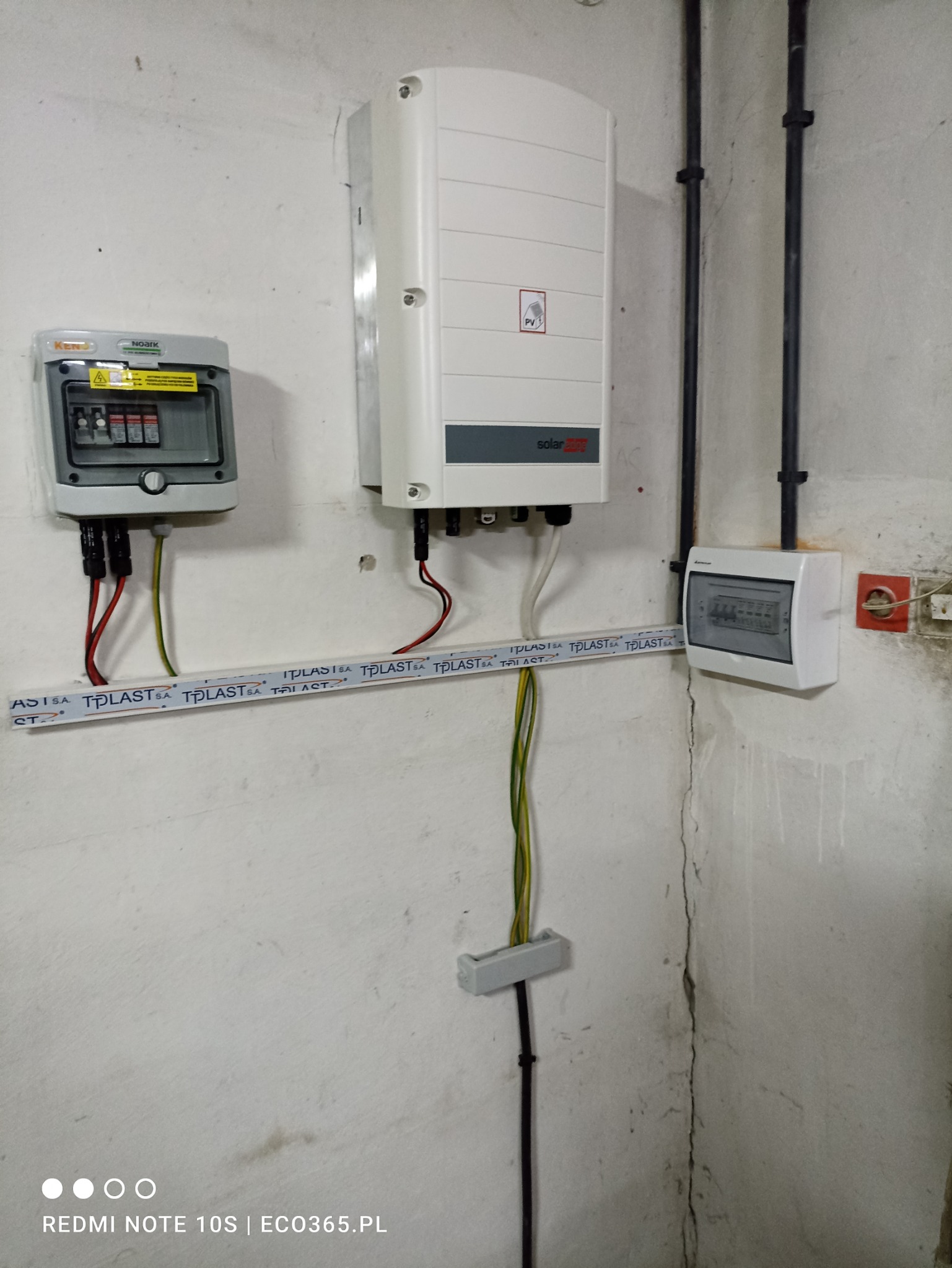 Biała obudowa falownika fotowoltaicznego SolarEdge z widocznym symbolem PV, zamontowana na szarej ścianie obok skrzynki elektrycznej z bezpiecznikami i przewodami w rurkach TPLAST.