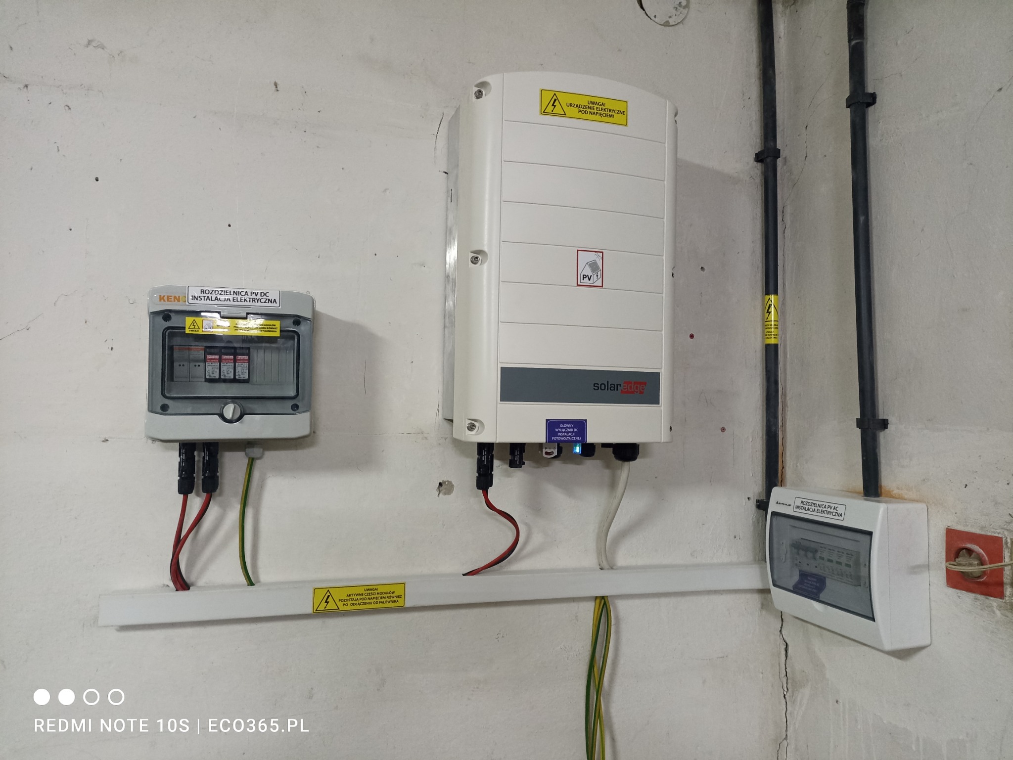 Instalacja fotowoltaiczna wewnątrz budynku: falownik SolarEdge, rozdzielnica PV DC firmy KEN i rozdzielnica PV AC, połączone przewodami na tle surowej, betonowej ściany.