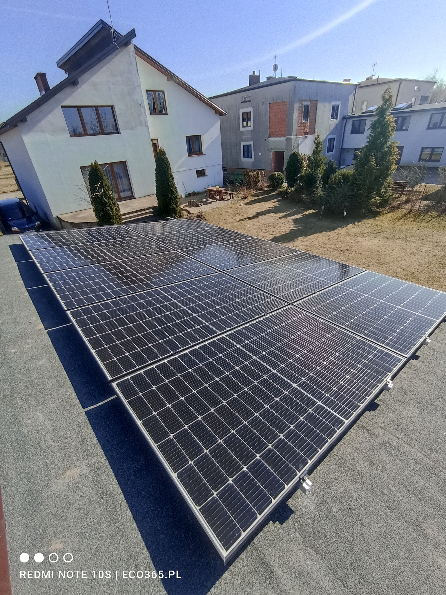 Panele słoneczne zamontowane na płaskim dachu budynku mieszkalnego, widoczne domy w tle, słoneczny dzień.