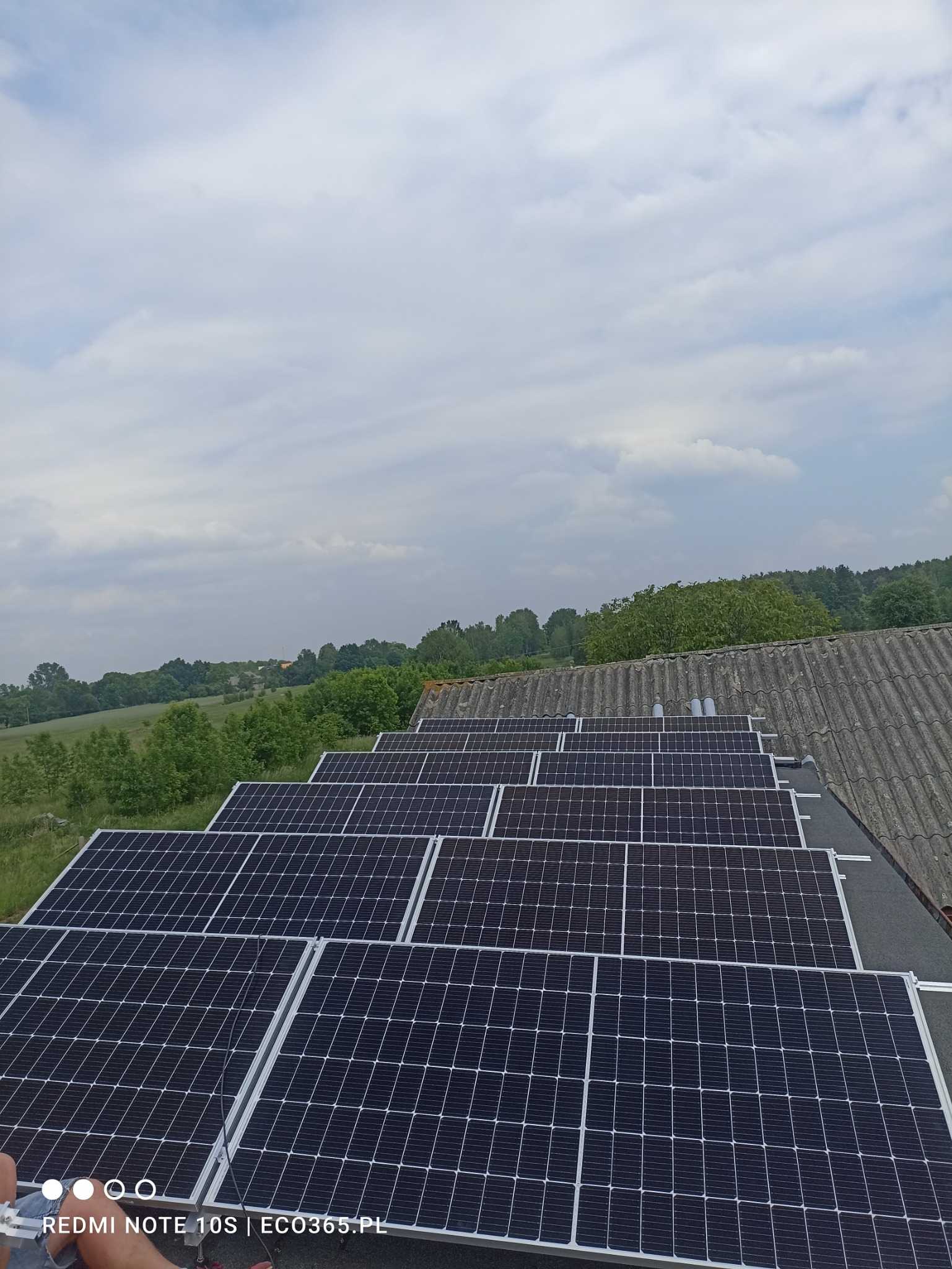 Rząd paneli słonecznych, z widocznym krajobrazem pól i lasów w tle, zamontowanych na dachu pokrytym szarą dachówką. Kąt ujęcia z dołu, ukazujący pochmurne niebo.
