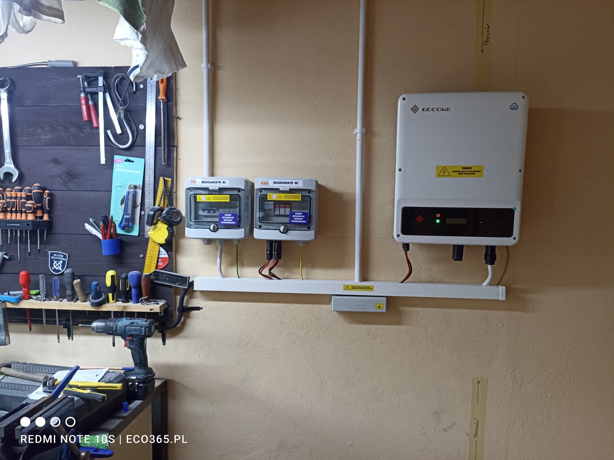 Zamontowany inwerter Goodwe z rozdzielnicami PV-AC i PV-DC na ścianie w warsztacie, widoczne narzędzia na tablicy i wiertarka na stole.