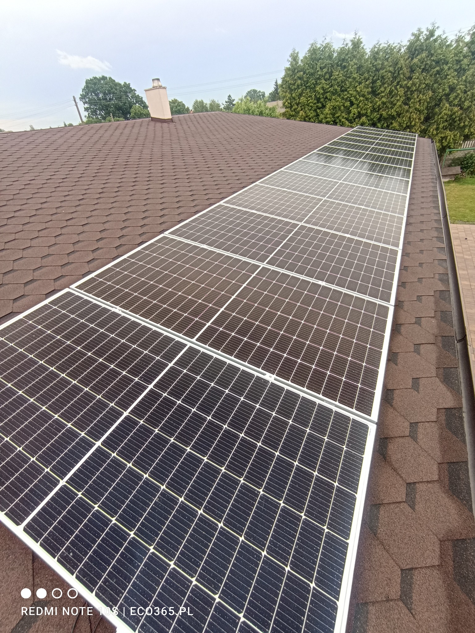 Panel słoneczny zamontowany na dachu pokrytym brązową dachówką bitumiczną, widok z góry na instalację.