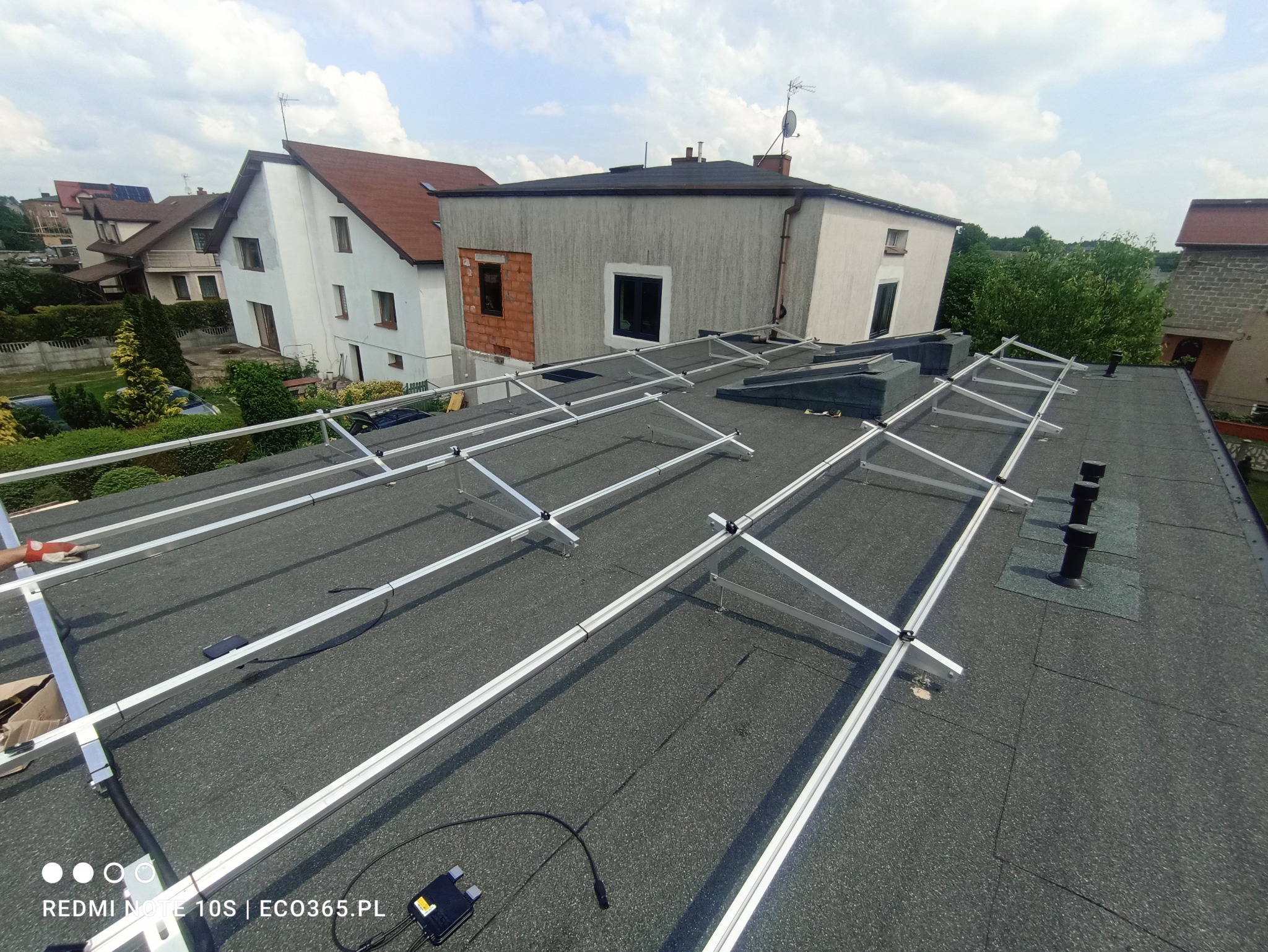 Montaż stelażu pod panele słoneczne na dachu pokrytym papą, widoczne kominki wentylacyjne i fragment ręki w czerwonej rękawicy.