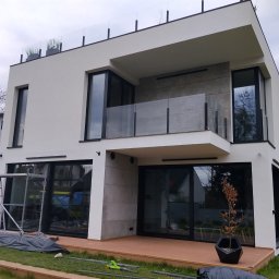 Givemo Michał Misiaszek - Nowoczesny, dwukondygnacyjny dom z białymi ścianami, dużymi oknami i balkonem z szklaną balustradą. Na pierwszym planie widoczne narzędzia i rusztowanie.