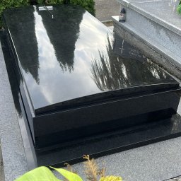 TOMBET NATALIA DOBIES - Czarny, błyszczący nagrobek z granitu, odbijający drzewa i niebo, z kwiatami w dolnym rogu, na tle cmentarza.