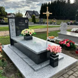 Nowoczesny, dwupoziomowy nagrobek z szarego i czarnego granitu z fotografią zmarłej, ozdobiony kompozycjami kwiatowymi w czarnych wazonach, na tle cmentarza z innymi grobami i drzewami.