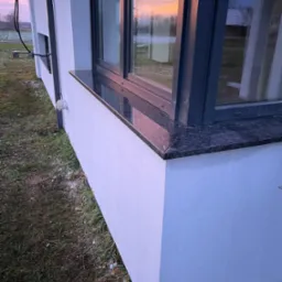 Zewnętrzny, ciemny, kamienny element pod oknem nowoczesnego budynku z białym tynkiem, widok na zachodzące słońce odbijające się w szybie.