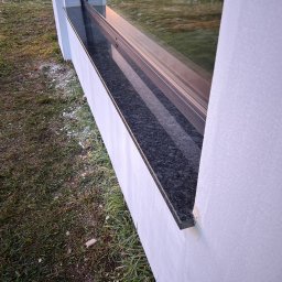 TOMBET NATALIA DOBIES - Zamontowany, ciemny parapet zewnętrzny z kamienia, widoczny z boku, z trawnikiem i resztkami śniegu w tle.