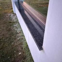 Zamontowany, ciemny parapet zewnętrzny z kamienia, widoczny z boku, z trawnikiem i resztkami śniegu w tle.