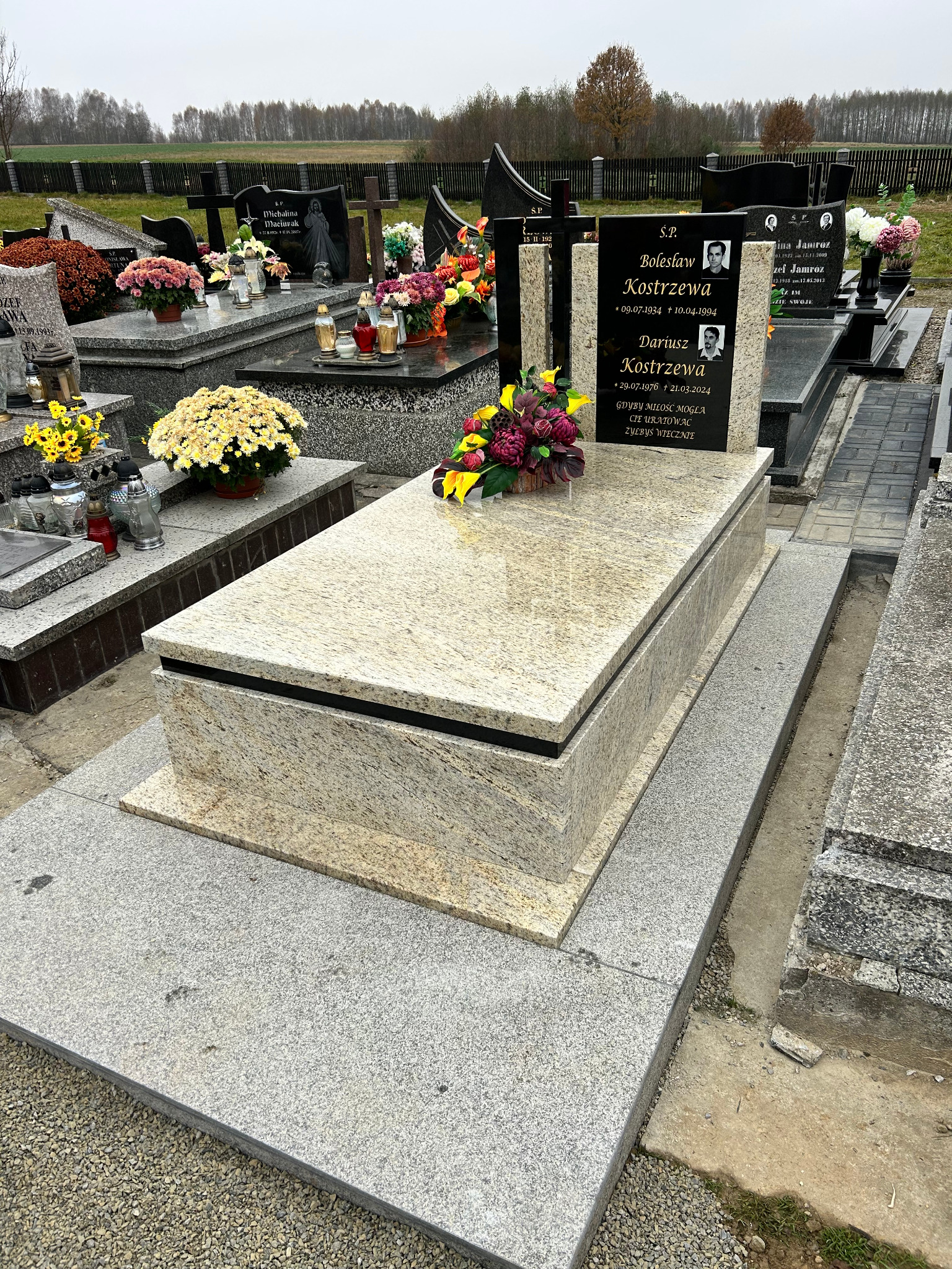 Jasny, granitowy nagrobek z czarną tablicą inskrypcyjną i zdjęciami zmarłych, udekorowany kwiatami, na cmentarzu.