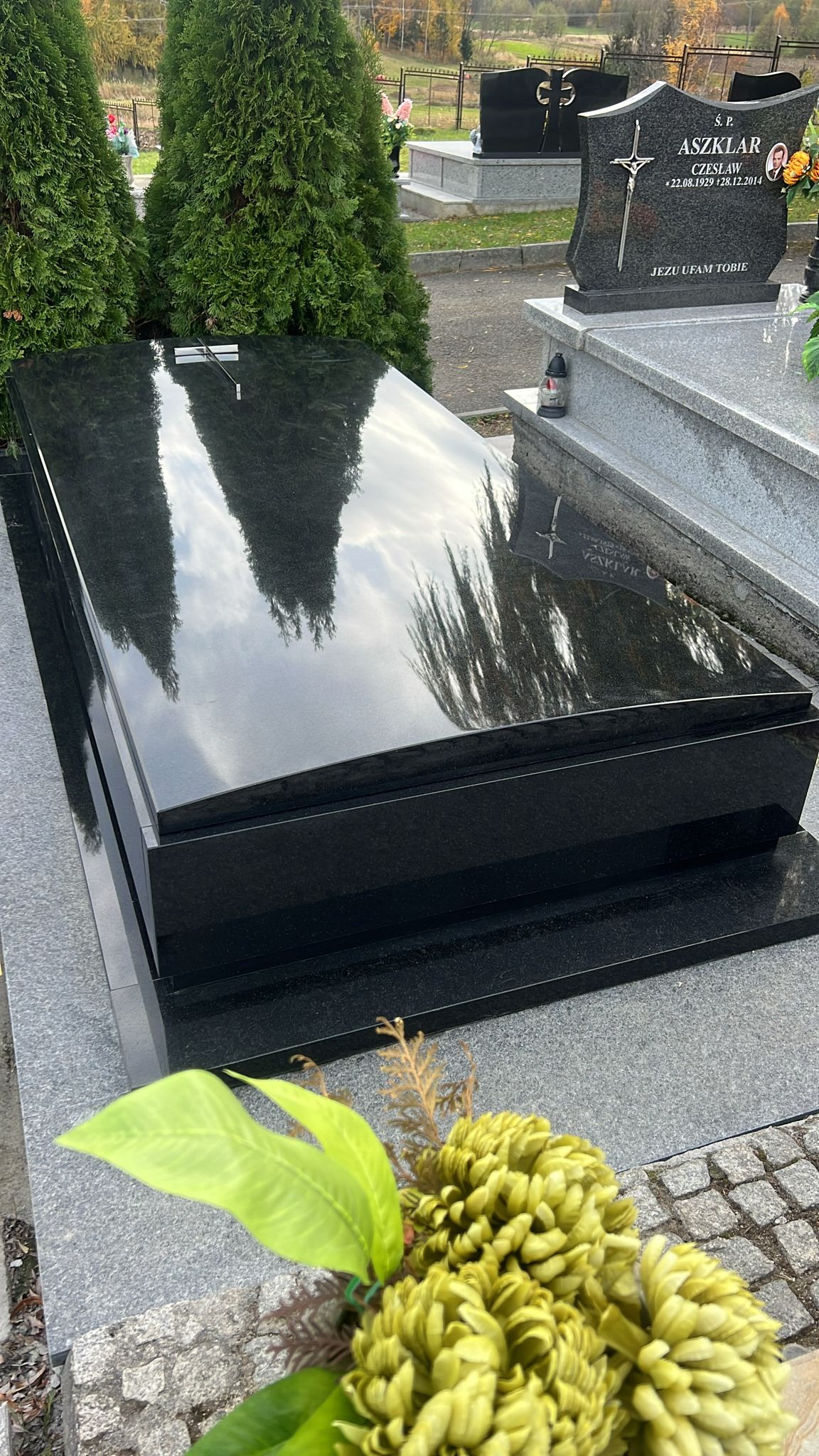 Czarny, błyszczący nagrobek z granitu, odbijający drzewa i niebo, z kwiatami w dolnym rogu, na tle cmentarza.