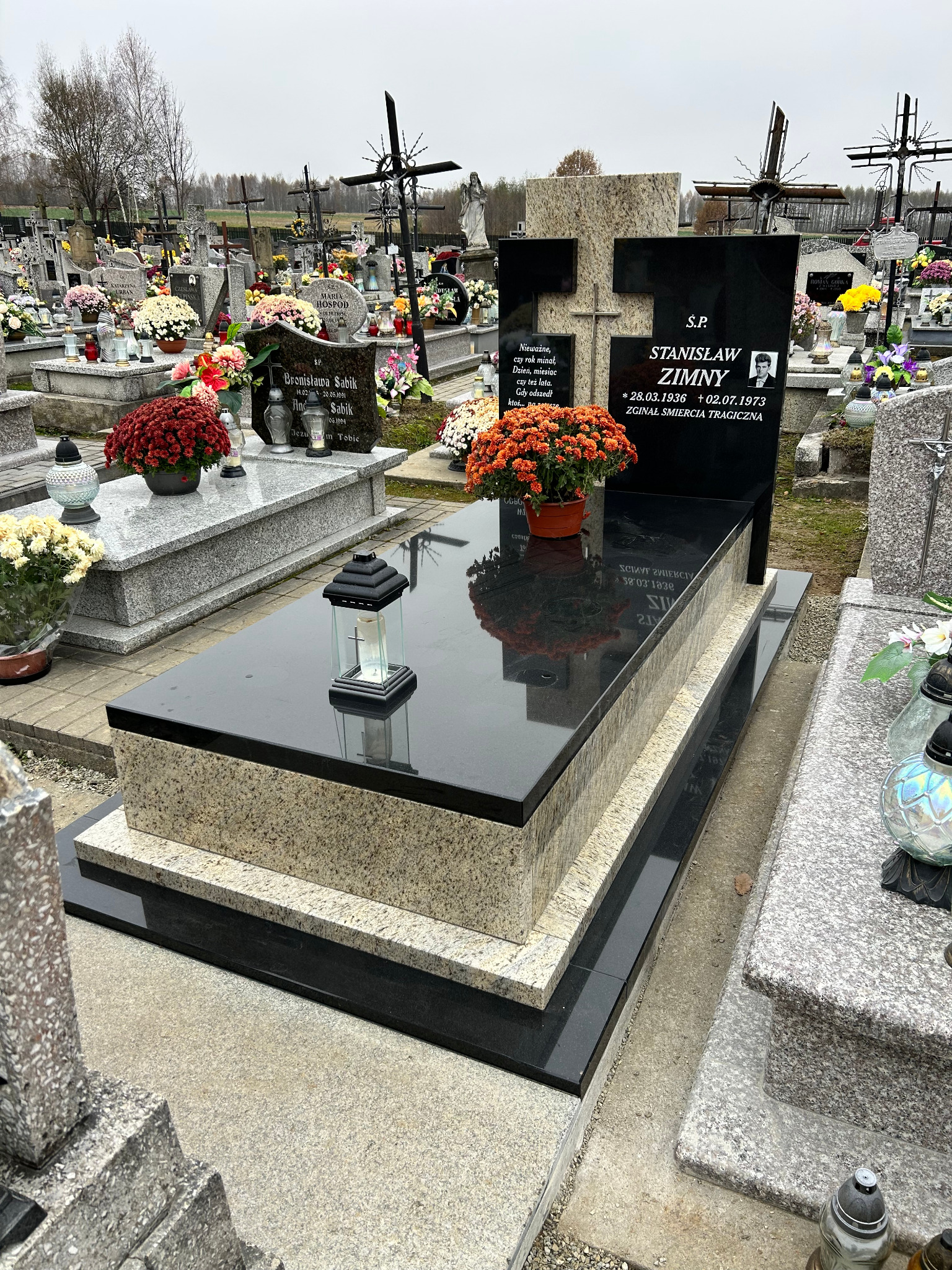 Czarny, polerowany nagrobek z jasnego granitu na cmentarzu, z pomarańczowymi chryzantemami w doniczce i lampionem na płycie.