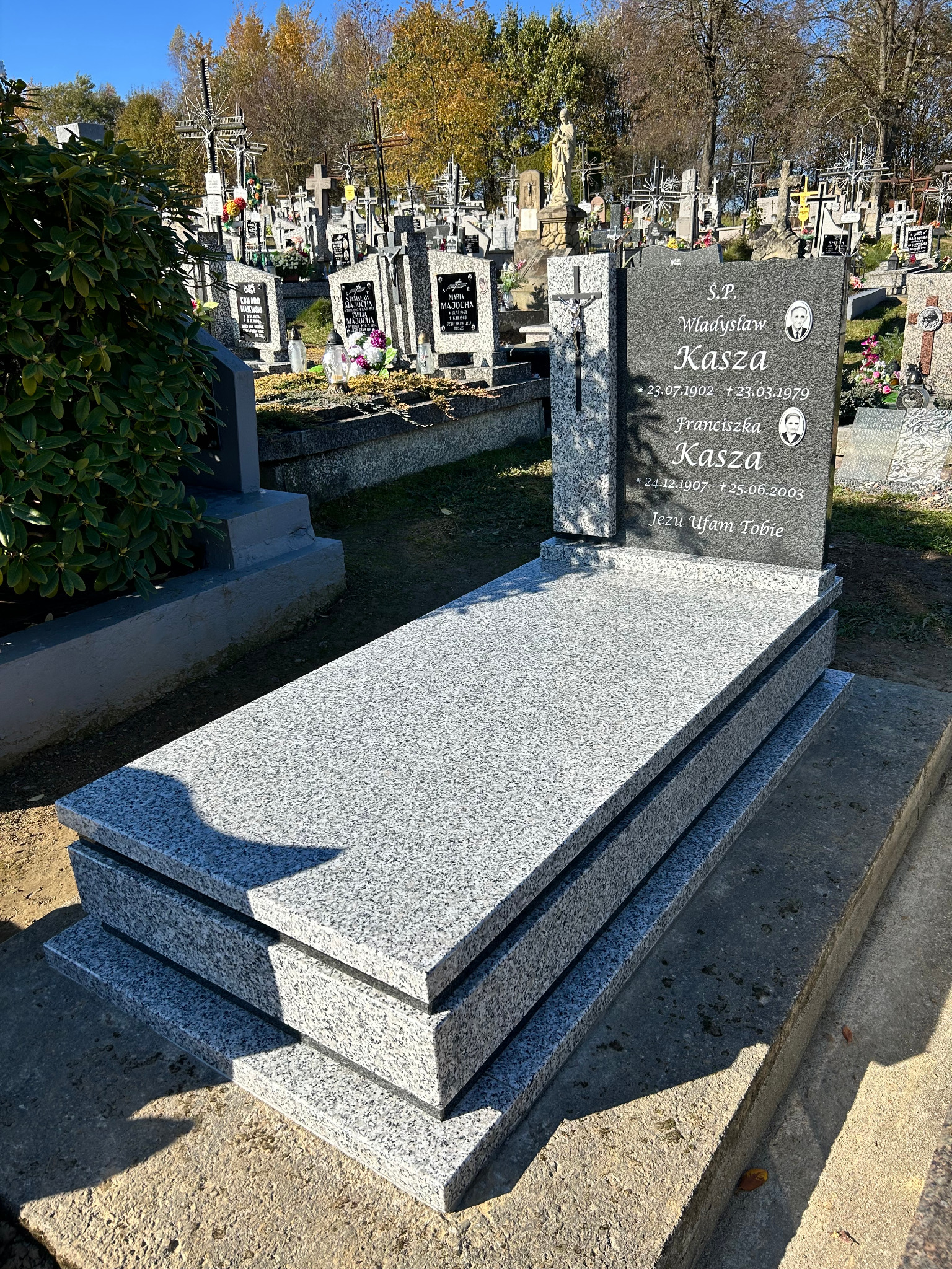 Granitowy nagrobek rodzinny z jasnoszarego kamienia na cmentarzu, z wygrawerowanymi danymi osób zmarłych i ich portretami.