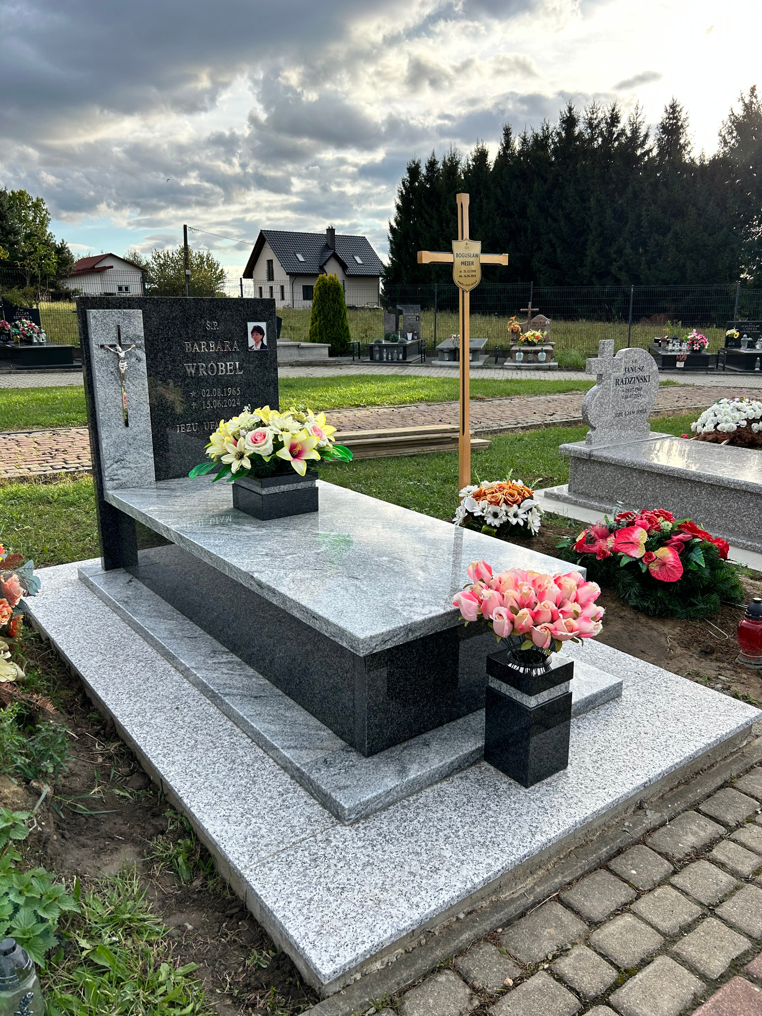 Nowoczesny, dwupoziomowy nagrobek z szarego i czarnego granitu z fotografią zmarłej, ozdobiony kompozycjami kwiatowymi w czarnych wazonach, na tle cmentarza z innymi grobami i drzewami.