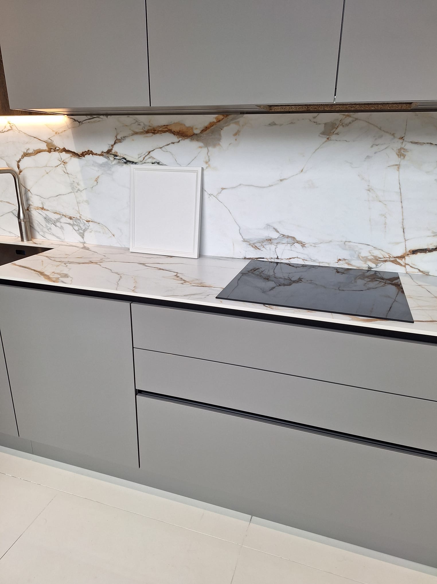 Szara zabudowa kuchenna z blatem imitującym marmur z wbudowaną płytą indukcyjną i oświetleniem LED podszafkowym, uzupełniona o pustą ramkę na zdjęcia.