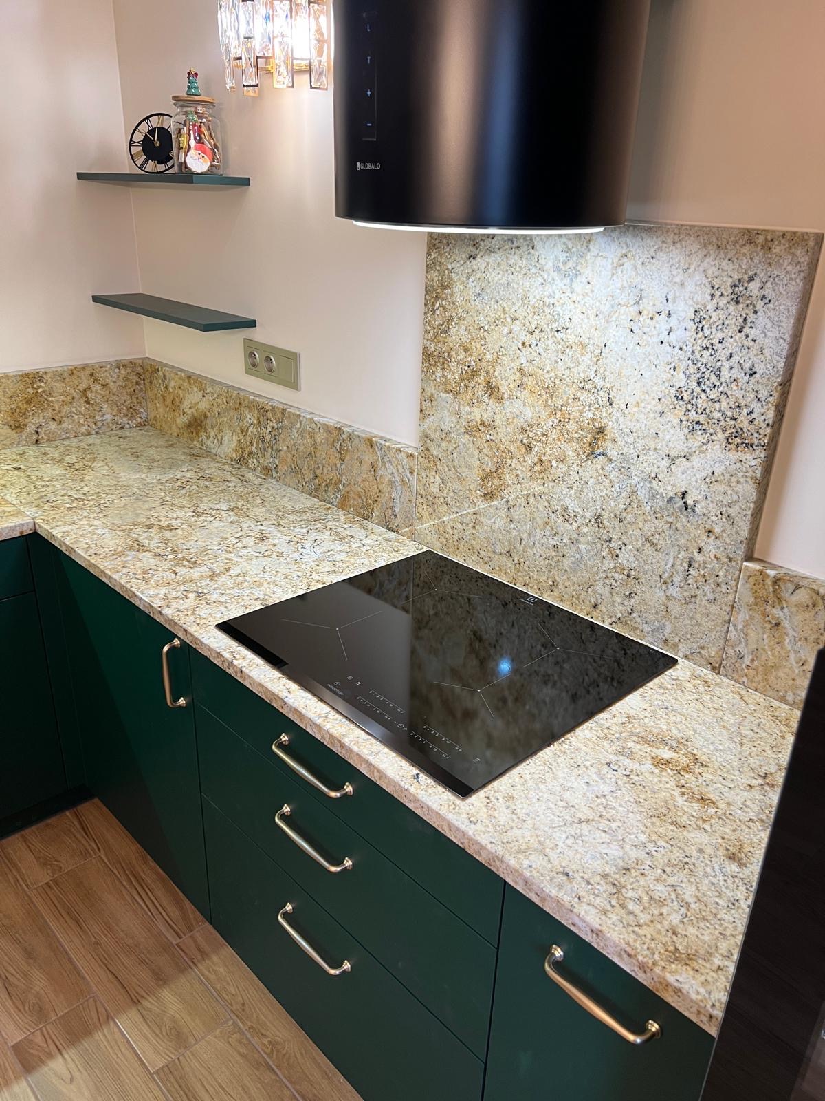 Kuchnia z zielonymi szafkami i blatem z kamienia o wzorze w odcieniach beżu i brązu, płyta indukcyjna wbudowana w blat, nad płytą czarny okrągły okap, na ścianie nad blatem panel z tego samego...