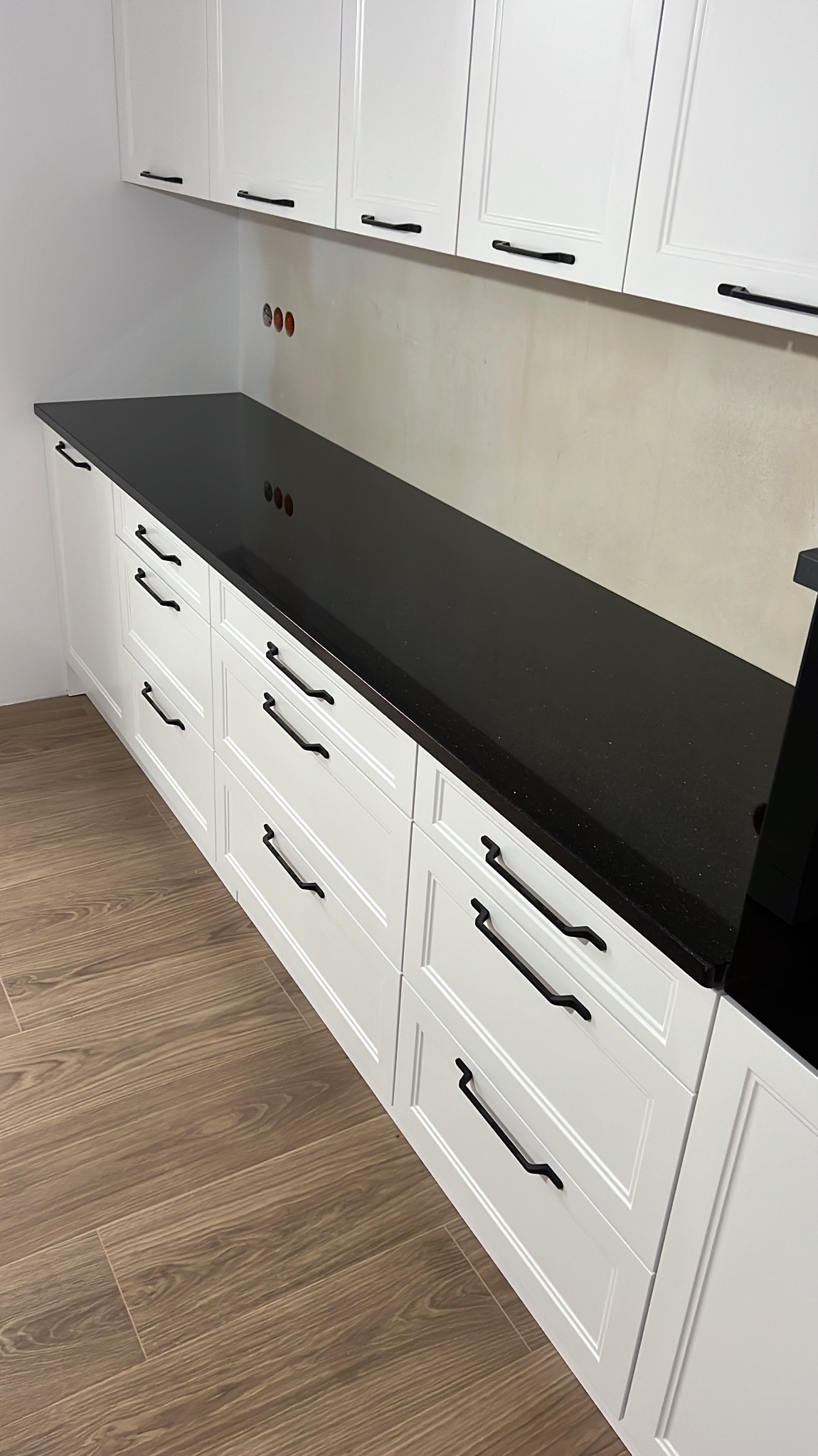 Długa, czarna powierzchnia robocza z drobnymi, jasnymi inkluzjami, zamontowana na białych szafkach kuchennych z czarnymi uchwytami, widoczne gniazdka elektryczne w ścianie.