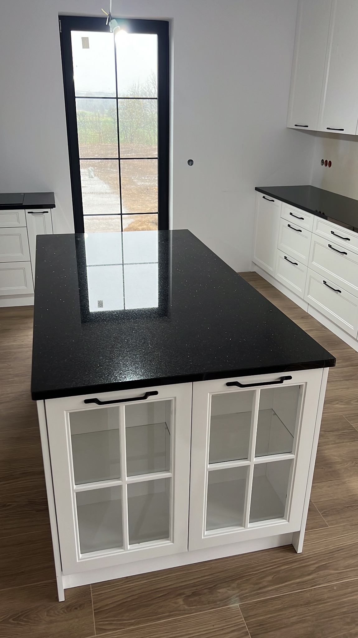 Wyspa kuchenna z czarnym blatem kamiennym z drobinkami, białe szafki z przeszklonymi drzwiczkami, czarne uchwyty, w tle okno z czarną ramą i białe szafki kuchenne.