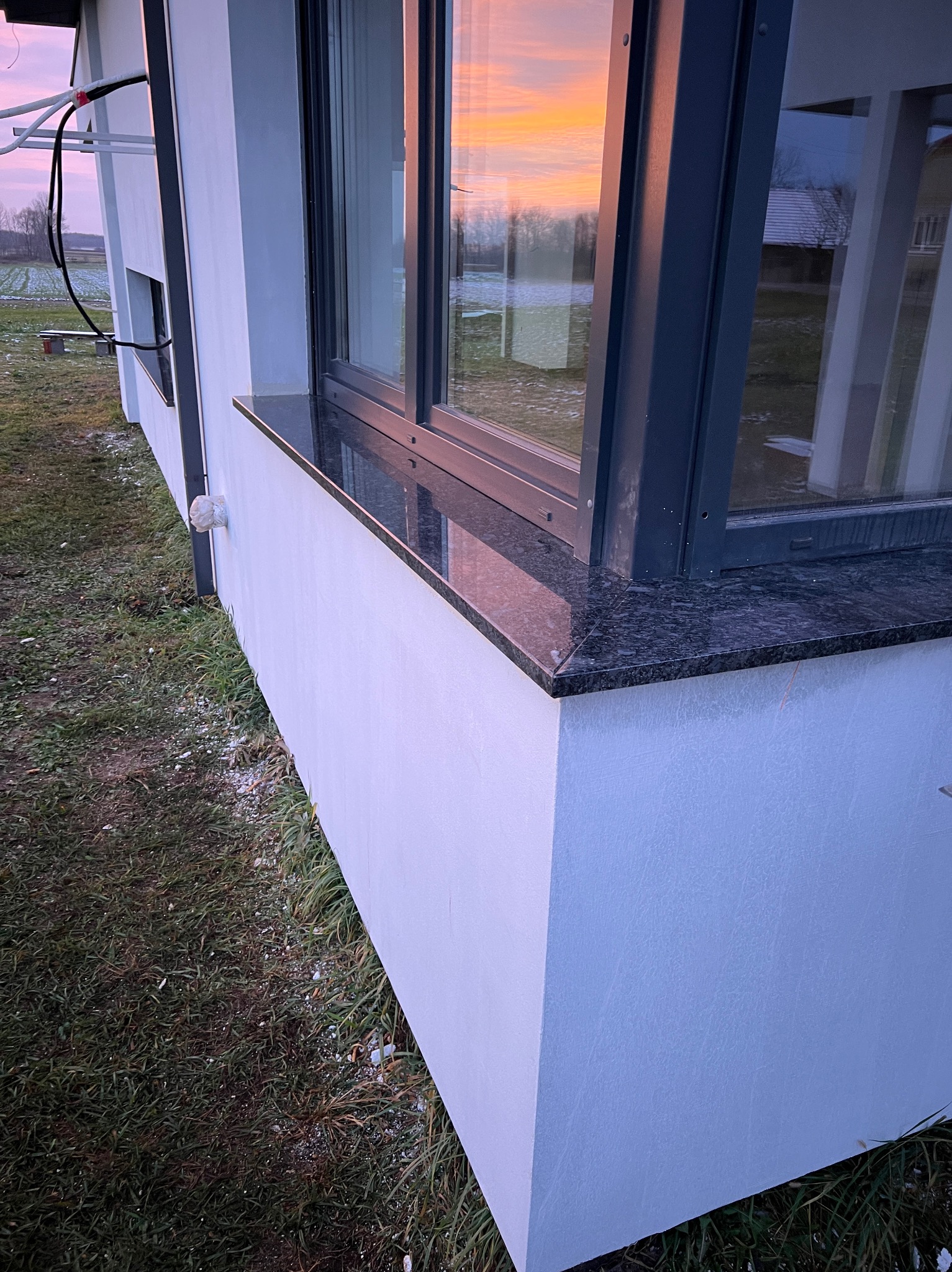 Zewnętrzny, ciemny, kamienny element pod oknem nowoczesnego budynku z białym tynkiem, widok na zachodzące słońce odbijające się w szybie.