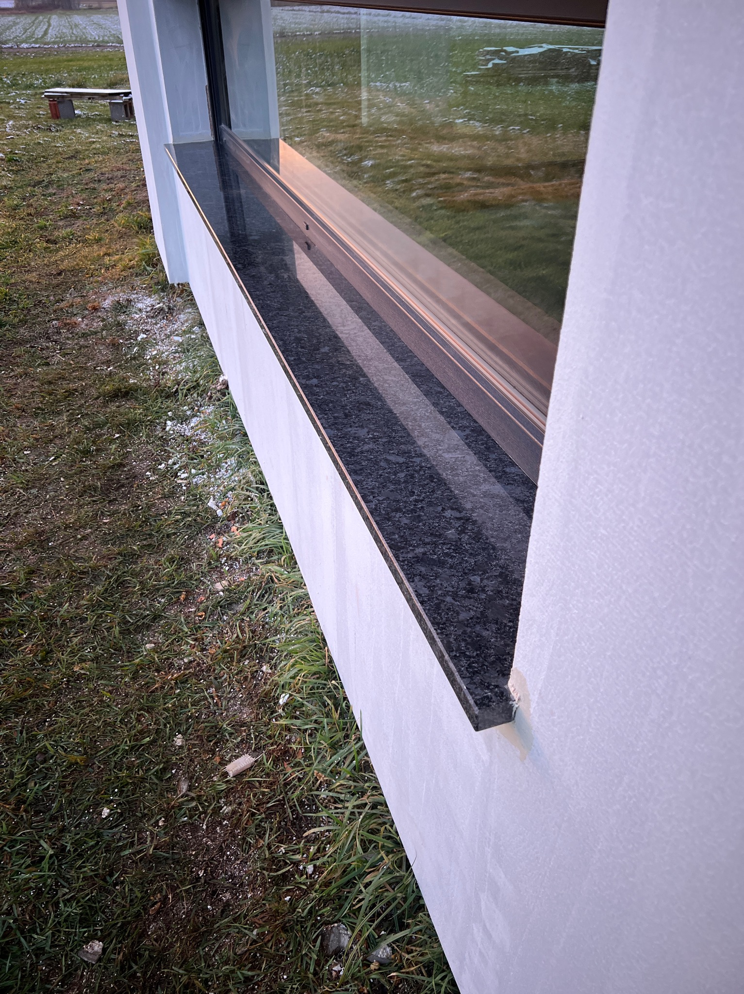 Zamontowany, ciemny parapet zewnętrzny z kamienia, widoczny z boku, z trawnikiem i resztkami śniegu w tle.