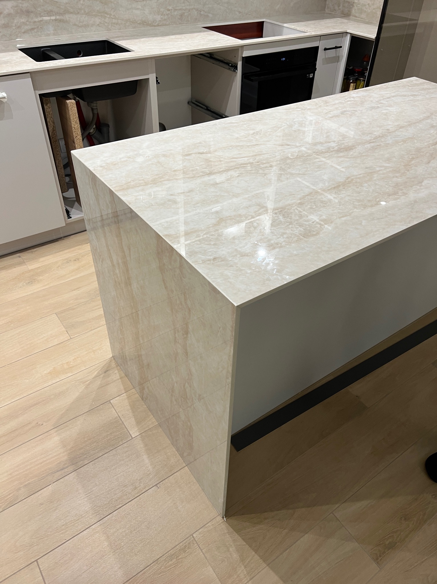 Beżowy blat kuchenny z wyspą, imitujący marmur, z widocznym zlewem i piekarnikiem w tle, na podłodze panele drewnopodobne.