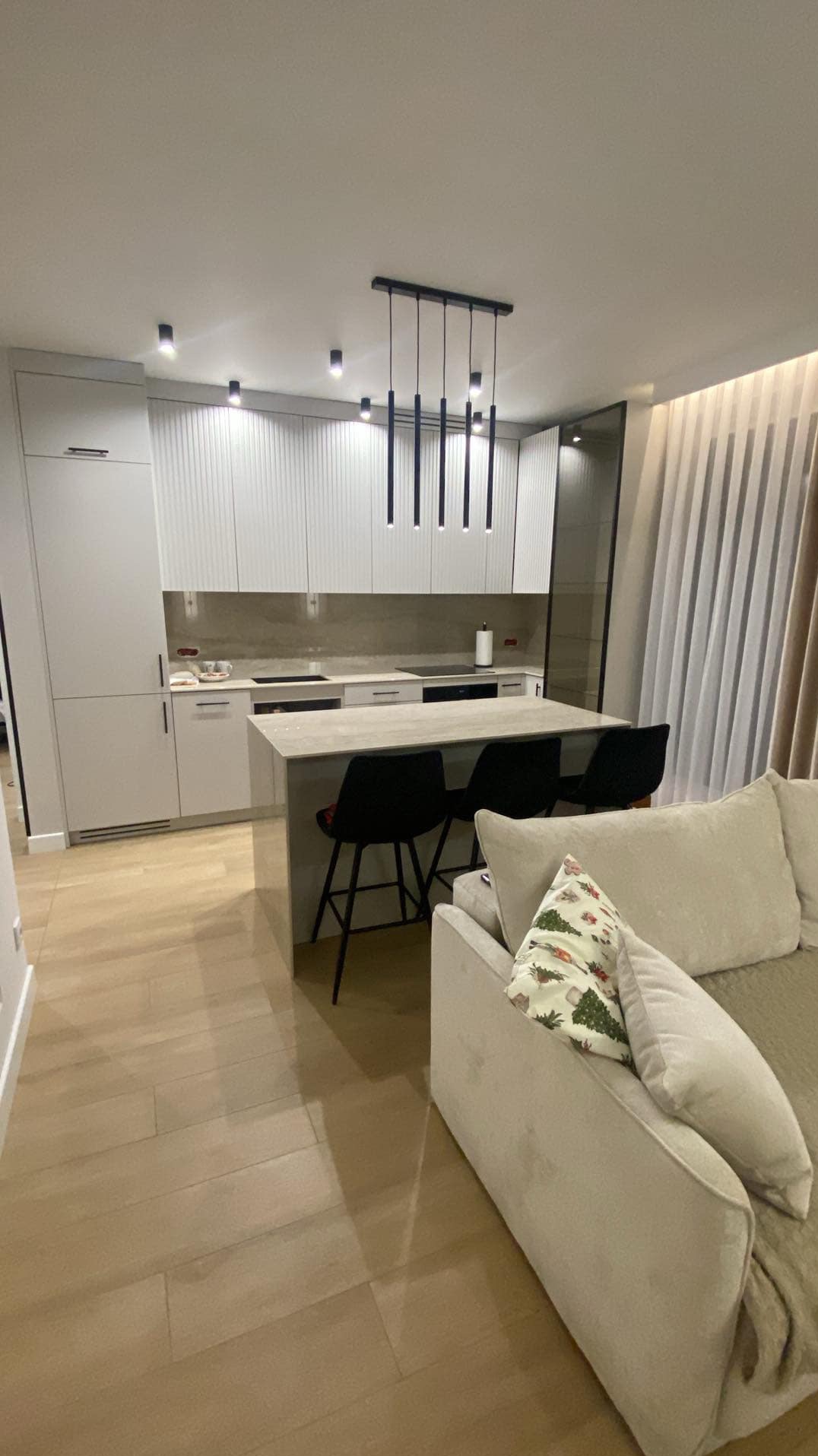 Nowoczesna kuchnia z wyspą i blatem z jasnego kamienia, widoczna z perspektywy salonu z beżową sofą i poduszką w świąteczne wzory.