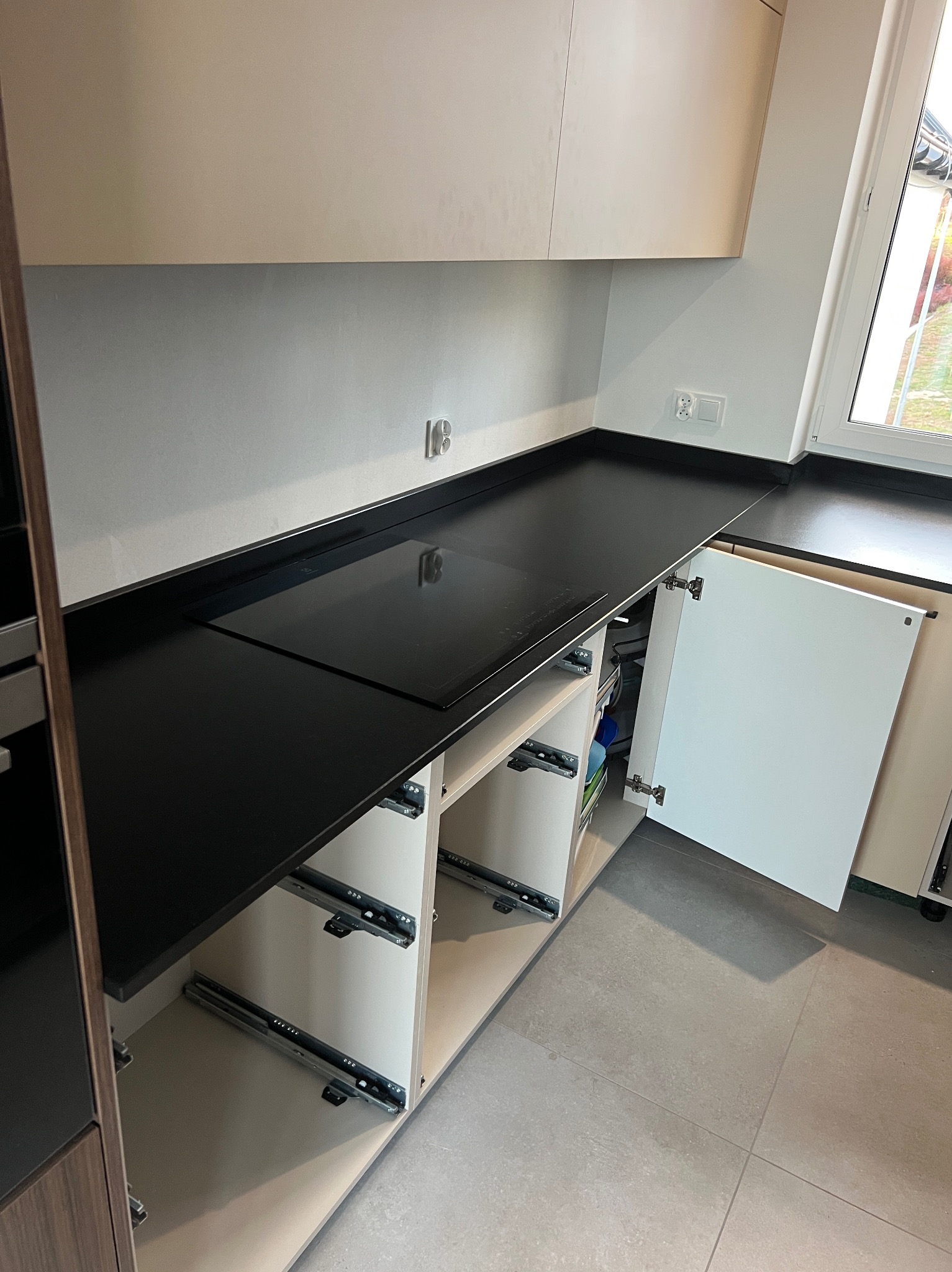 Czarny, błyszczący blat kuchenny z wbudowaną płytą indukcyjną, widoczne szuflady z systemem cichego domykania i częściowo otwarte białe drzwiczki szafki w nowoczesnej kuchni.