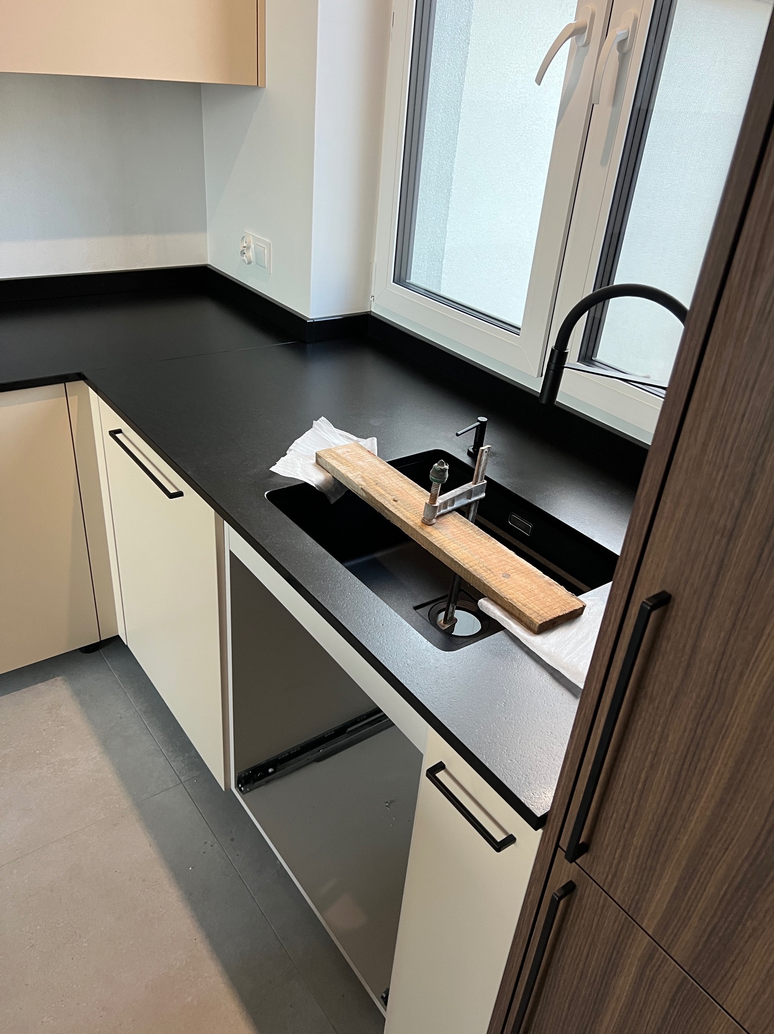 Czarny blat kuchenny z zamontowanym zlewem, widoczny fragment okna i szafek kuchennych, w zlewie drewniana deska mocowana ściskiem stolarskim.