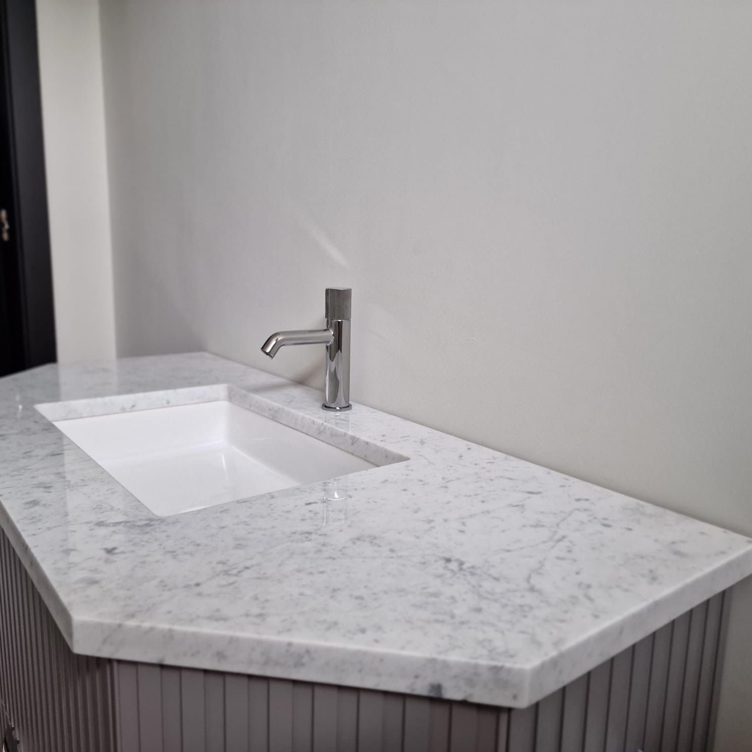 Biały marmurowy blat kamienny z wpuszczoną umywalką i chromowaną baterią, widoczny fragment szafki z pionowymi żłobieniami.