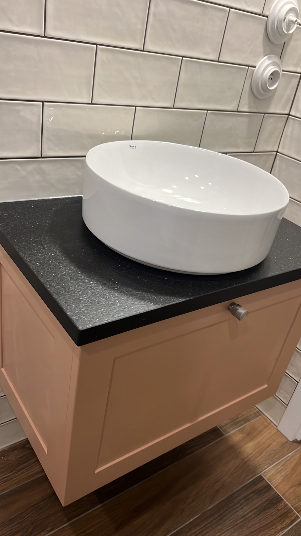 Okrągła, biała umywalka nablatowa marki Roca na czarnym blacie z drobinkami, umieszczona na szafce w kolorze pudrowego różu, z widocznymi płytkami ceramicznymi na ścianie i drewnopodobną podłogą.