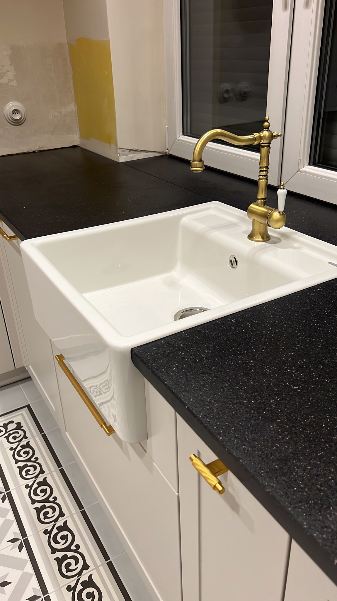 Kuchnia z czarnym blatem nakrapianym, białym zlewem farmerskim, baterią w stylu retro z mosiądzu i białymi szafkami z złotymi uchwytami.