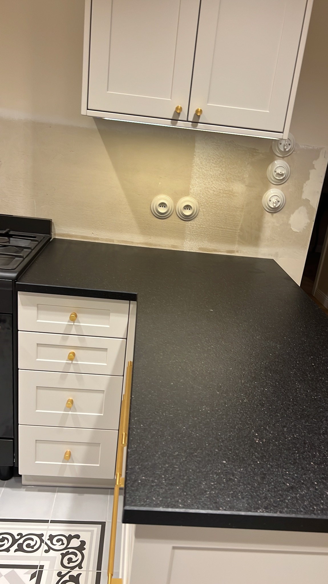 Czarny, błyszczący blat w kuchni z białymi szafkami i złotymi uchwytami, widoczne gniazdka elektryczne na ścianie w tle.