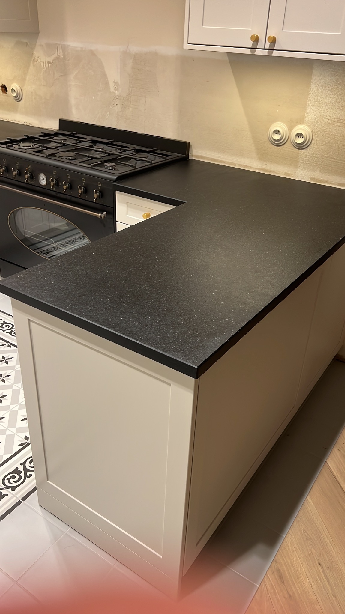 Czarny, matowy blat kuchenny z delikatnym wzorem, zamontowany na beżowej wyspie, z widoczną kuchenką retro w tle i fragmentem wzorzystej podłogi.