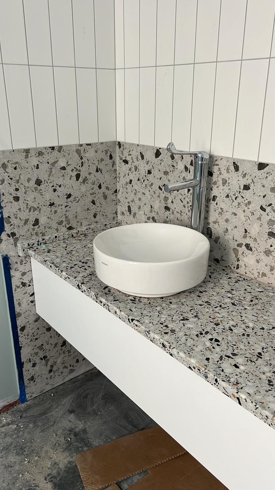 Łazienkowa umywalka nablatowa na blacie z lastryko, biała szafka pod umywalką, chromowana bateria, białe pionowe płytki na ścianie, fragment niezabezpieczonej podłogi w tle.